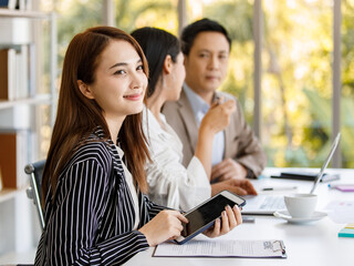 young attractive Asian businesswoman dressed formally sitting and holding tablet with smiley face during working in the office with colleagues sitting in the background