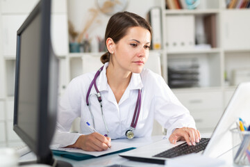 Experienced female physician filling up medical forms on laptop while sitting at table in office