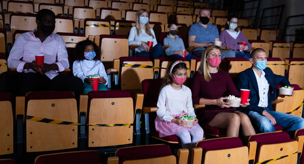 Men, women and children in protective face masks sitting in cinema auditorium. Coronavirus pandemic safety rules, social distance during movie watching