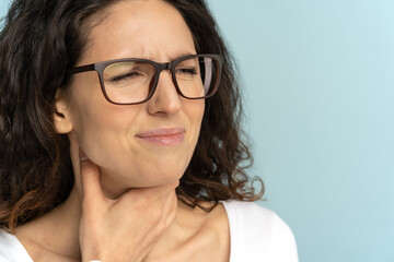 Closeup of sick woman wear glasses having sore throat, tonsillitis, caught cold, suffering from painful swallowing, strong pain in throat, holding hand on her neck, isolated on studio blue background