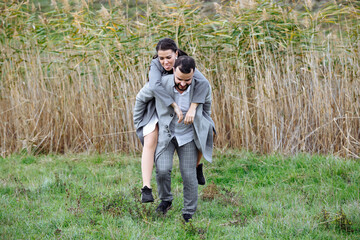 Naklejka na ściany i meble A stylish couple in gray coats are having fun. A man holds a woman on his back and she hugs him.
