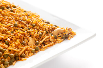 Hot spicy Kashmiri Mixture (Namkeen) snacks in a white ceramic square plate made with dry fruits, peanuts, corn flakes, etc. Pile of Indian spicy snacks, under backlight, side view
