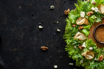 Obraz na płótnie Canvas Light diet salad with pear, nuts and blue cheese. The salad is laid in the form of a wreath on a round wooden board with sauce in the center on a dark background. Copy space Top view