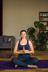female yogi in sportswear sits in a lotus position on a yoga mat in a room. Yoga at home