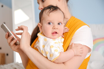 Young mother with little baby in sling using mobile phone at home