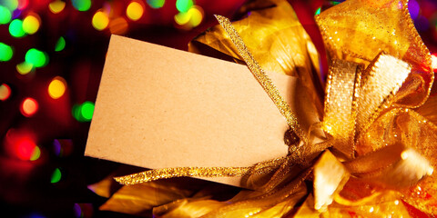 Gold bow on a gift box with a label for the text, multicolored lights garlands on the background, close-up, top view.