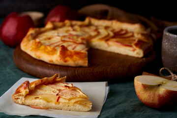 Apple pie with piece slice of galette with fruits, sliced sweet pastries on dark green tablecloth, sweet crostata on cutting wooden board, side view, autumn or winter food