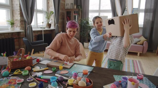 Zoom in shot of happy little boy putting on Easter bunny box on head with help of cheerful mother and sister and then jumping and playing at home
