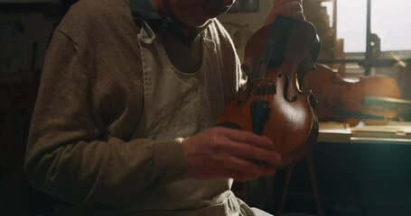 Cinematic shot of experienced master artisan luthier controlling quality of handmade wood violin just made in creative workshop. Concept of spiritual instrument, art, orchestra, passion for music.