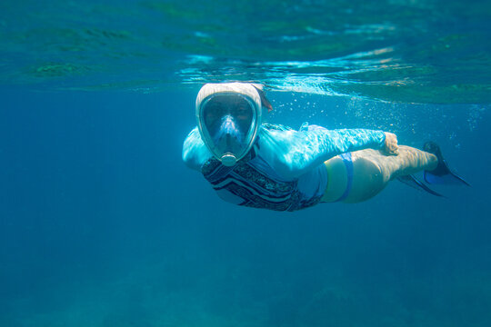 Woman swimming in blue water in full-face mask. Young woman swimming on sea surface. Snorkel in coral reef of tropical sea. Woman in snorkeling mask underwater photo. Blue water sport activity.