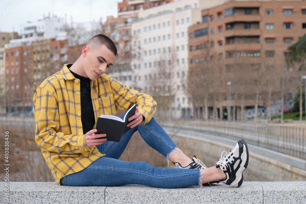 Wall mural Handsome young man wearing make up, smiling and reading a book sitting on a wall. Non binary androgynous guy.