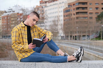 Handsome young man wearing make up, smiling and reading a book sitting on a wall. Non binary androgynous guy.