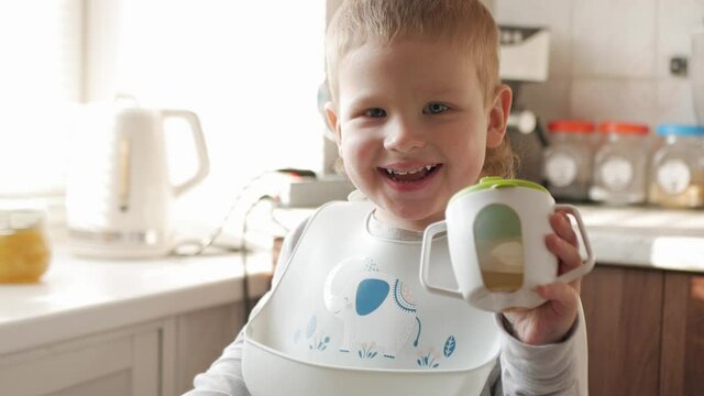 A boy in a bib and a highchair sits at the kitchen table. The child looks at the camera, drinks peach juice from a sippy cup, smiles and enjoys the process.