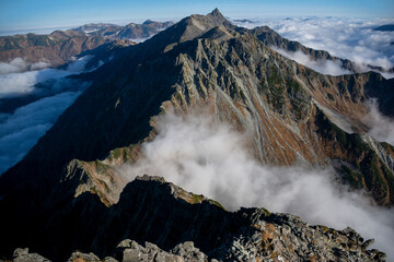 信州、北アルプス、穂高連峰、北穂高岳（標高3,106m ）山頂の道標と雲海に浮かぶ槍ヶ岳、大キレット、南岳、大喰岳。青空。	