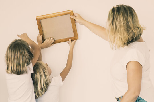 Blonde mom and daughters hanging blank photo frame on white wall. Back view of Caucasian mother holding and aligning empty picture with help of two girls. Family, relocation and moving day concept