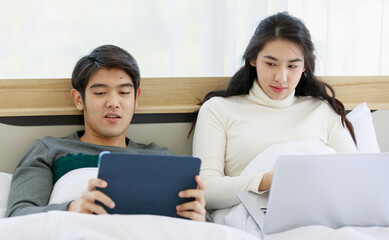 Portrait of cute smiling young Asian lover couple in white and gray long sleeve sweatshirt lying on a bed while using a dark blue digital tablet together. Woman see funny things and laugh out