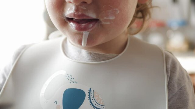 Close-up of a child's grimy mouth eating dairy products yogurt, sour cream, cottage cheese. A boy in a bib and a highchair sits at the kitchen table.