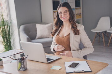 Photo portrait of pregnant lady touching belly sitting at desk in modern office