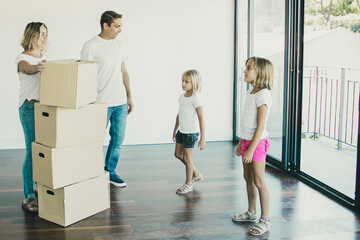 Fototapeta na wymiar Caucasian standing in room with balcony and looking at each other. Happy holding stack of carton boxes and smiling. Father talking with girls. Mortgage, relocation and moving day concept