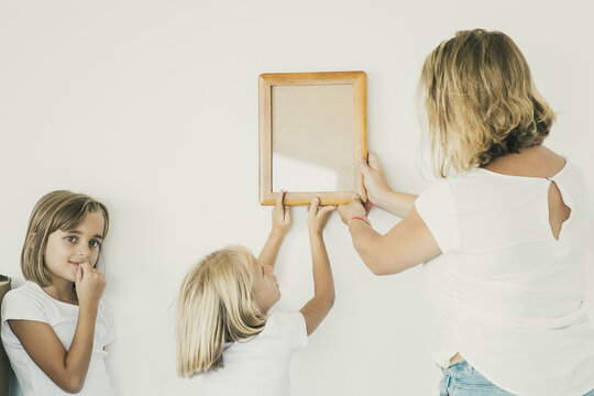 Lovely girl putting frame on white wall with help of mom. Cute daughter hanging blank picture. Her sister standing near and looking away. Family decorating room. Relocation and moving day concept