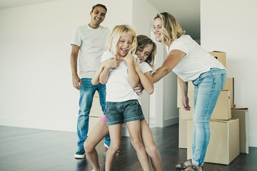 Joyful family couple and two kids having fun while moving into new apartment. Girls tickling each other and laughing. Full length. Real estate purchase concept