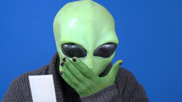 Humanoid in cozy knitted jacket looks at photo, postcard or reads note, blue background. Sick alien sneezes or coughs. Actor plays in science fiction movie or theater production.