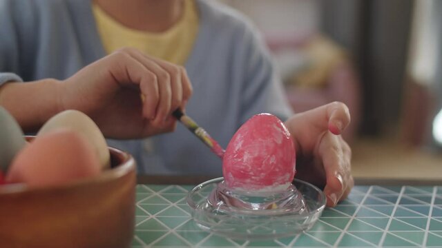 Close up shot of hands of little girl drawing with pink paint on egg in glass holder while preparing for Easter holiday