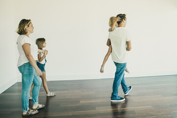Fototapeta premium Side view of dad holding daughter and going to balcony with her. Pretty blonde mother in casual clothes standing with another daughter, holding her hand and smiling. Relocation and moving day concept