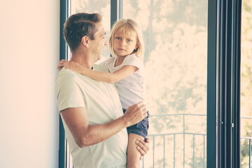 Cheerful father holding cute daughter, looking at her and smiling. Lovely blonde girl looking at camera. Happy dad with kid standing near balcony open door. Relocation and family moving day concept