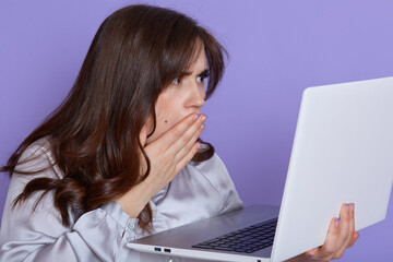 Amazed emotional woman with dark hair, looking at laptop screen with shocked expression, covering her opened mouth, isolated over lilac background, sees something bad, has trouble with work.