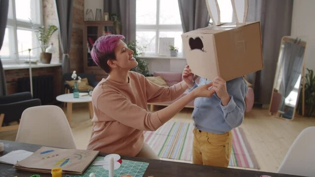 Zoom in shot of cheerful mother putting Easter bunny box on head of little daughter, sticking paper nose with teeth on mask and laughing while watching how girl dancing