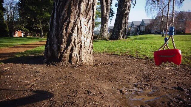 Moving Lonely Red Plastic Swing Hangs Above Mud On Big Tree In A Beautiful Green But Empty Park In UK During The Covid 19 Pandemic. 