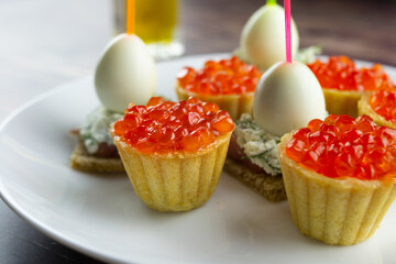 canapes with red caviar, salmon and quail eggs on a white plate, tartlets, small appetizing sandwiches on skewers, bread with cream cheese on a wooden background