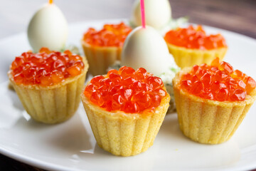 canapes with red caviar, salmon and quail eggs on a white plate, tartlets, small appetizing sandwiches on skewers, bread with cream cheese on a wooden background