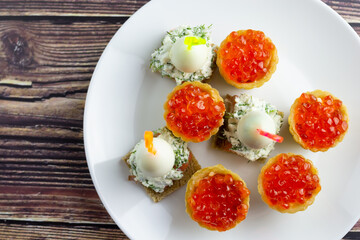 canapes with red caviar, salmon and quail eggs on a white plate, tartlets, small appetizing sandwiches on skewers, bread with cream cheese on a wooden background