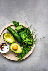 Rohes grünes Gemüse und eine halbierte Avocado auf einem Holz Teller. Grauer rustikaler Tisch, Flat lay, gesunde Ernährung.