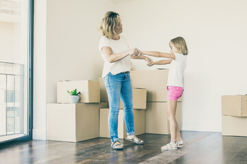 Loving mother having fun and dancing with preschooler daughter in new home. Happy Caucasian mom playing with two girls in room with carton boxes. Family, relocation and moving day concept