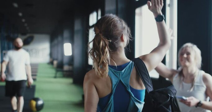 Camera follows young athletic blonde woman with backpack entering large modern gym, greeting friends before working out.
