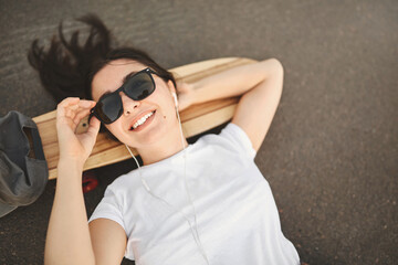 Urban lifestyle and skateboarding concept. Upper view smiling girl in sunglasses lying on concrete and penny board, hand behind head, gaze sunbeams, riding at skatepark, listen music in earphones