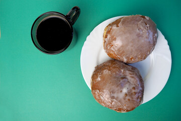 two sugar coated doughnuts lie in a white round plate next to a mug of hot custard coffee on a green background top view