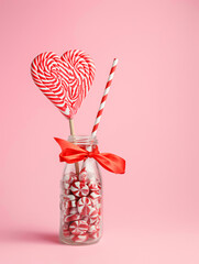 Jar full of sweet candies with heart shaped swirl lollipop and straw on pink background. Minimal Valentines day, romance or wedding concept. Flat lay.