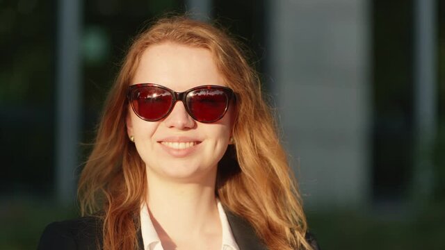 [4k] beautiful blonde business woman with sunglasses smiling at camera in golden sunlight with park reflecting in sunglasses