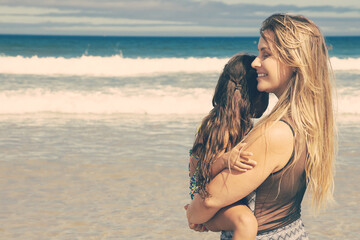 Happy relaxed young mom holding little daughter in arms, carrying kid, standing on beach. Bright ocean with waves and white foam, clear sky in background. Parenting and outdoor activities concept