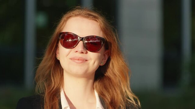 [4k] a young beautiful blonde business woman with sunglasses and reflections of a park und the sun on her glasses smiling at camera sorting her hair in golden sunlight