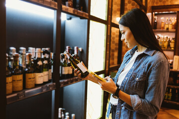 Beautiful young brunette woman buying wine examines label holding bottle