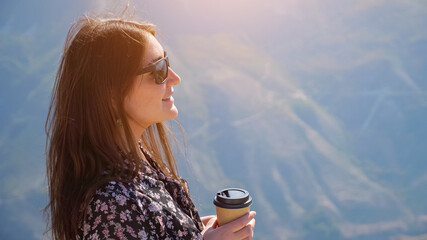Brunette woman with stylish sunglasses in color dress drinks coffee enjoying wonderful landscape of...