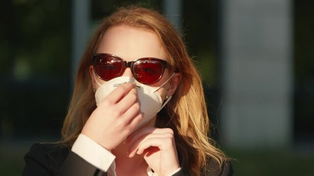 [4k] a beautiful young blonde business woman wearing sunglasses putting on a face mask vor covid-19 protection in golden sunlight smiling