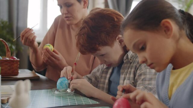 Side view of cute little brother and sister drawing with paint on Easter eggs while preparing for holiday with cheerful mother