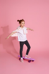 Stylish little child girl with skateboard in casual clothes riding skateboard over pink background.