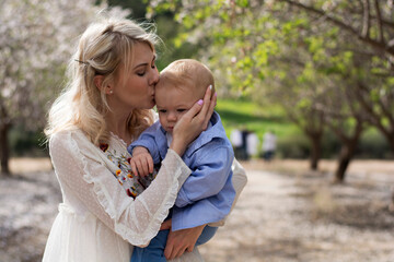 Fototapeta na wymiar Pretty woman holds in her arms boy. Mom gently hugs and kisses her son. Feeling of tenderness, love, care and joy. Family values for life. Spring is wonderful period of flowering almonds in nature.